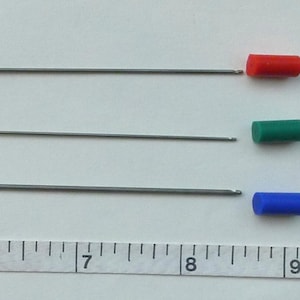 May include: Three metal rods with black, red, green, and blue coloured caps on the ends. The rods are approximately 20 cm long and are lined up horizontally on a white surface with a ruler underneath.