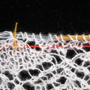 May include: Close-up of a white crocheted lace fabric with a red and blue thread running through it. A wooden knitting needle and a yellow knitting needle are visible.