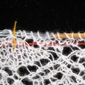 May include: Close-up of a white crocheted lace fabric with a red and blue thread running through it. A wooden knitting needle is resting on top of the fabric.