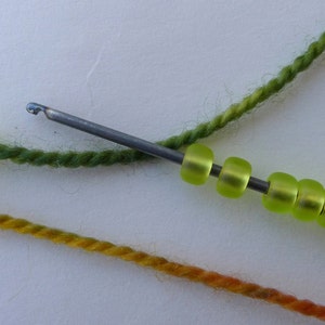 May include: A close-up of a crochet hook with a green yarn and a string of green beads. The beads are strung on the hook and the yarn is wrapped around the hook.