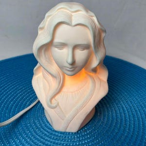 Peut inclure: Lampe buste en céramique blanche représentant une femme aux cheveux longs et ondulés. La lampe émet une lueur douce et chaleureuse. Le buste est posé sur une surface bleue, avec un cordon électrique blanc.