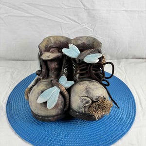 Peut inclure: Une paire de bottes décoratives marron patinées avec des accents de libellules bleu clair. Les bottes ont des lacets marron foncé et sont posées sur un tapis circulaire bleu. Aspect rustique et vieilli.