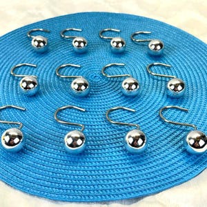 May include: A set of twelve silver-colored shower curtain hooks with a ball design. The hooks are arranged on a blue, circular, textured surface. The hooks have a curved top for hanging.