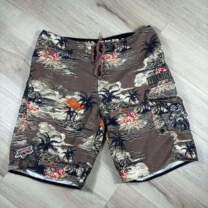 May include: Brown board shorts with a tropical print featuring palm trees, flowers, and a sunset scene. The shorts have a drawstring closure and a cargo pocket with the text "FOXRIDE". A Fox Racing logo is visible.