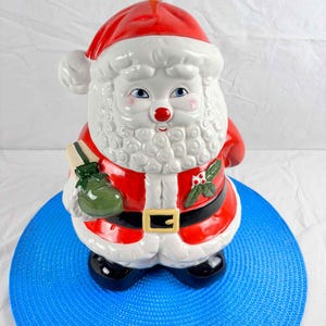 Pode incluir: Uma figura de cerâmica do Pai Natal com chapéu e fato vermelhos, barba branca e botas pretas. A figura segura um saco de presente verde e tem um cinto preto com fivela dourada. A figura do Pai Natal tem aproximadamente 33 cm de altura.
