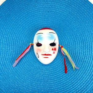 Dekorative Vintage-Gesichtsmaske aus Keramik mit stilisiertem, sanftem Ausdruck und bunten Bändern im Retro-Kunststil.