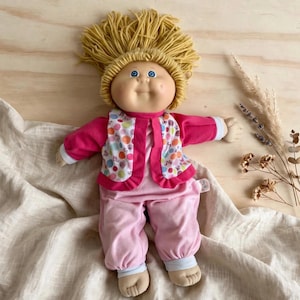 Puede incluir: Una muñeca con cabello de lana rubio y ojos azules, que viste una camisa rosa de manga larga, pantalones rosas y un chaleco blanco con un patrón de lunares de colores. La muñeca tiene un rostro suave y redondeado con dedos visibles.