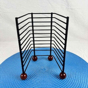 May include: A black metal wire rack with a geometric design. The rack has a rectangular shape with horizontal bars and is supported by three small, round, dark red feet. The rack is on a blue textured surface.