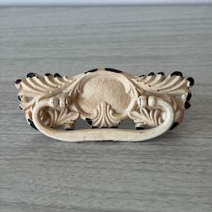 Op de afbeelding: Een decoratieve ladeknop met een gebroken witte afwerking en versleten zwarte randen. Het beslag heeft een sierlijk, symmetrisch ontwerp met bladmotieven, een centraal rond element en een gebogen, scharnierende handgreep.