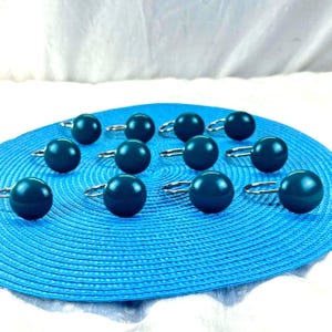 May include: A collection of teal-colored, spherical curtain tiebacks with silver-tone metal loops. The tiebacks are arranged on a textured blue circular mat, with a white background.