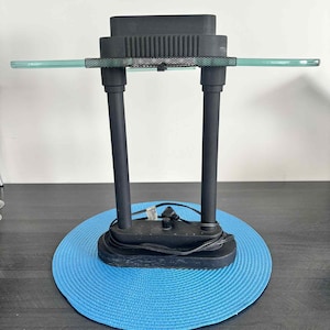 Peut inclure: Une lampe de bureau noire avec un dessus rectangulaire et deux supports verticaux. La lampe a une étagère en verre transparent et repose sur un tapis circulaire bleu. La lampe est branchée et prête à l'emploi.