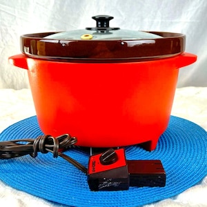 Könnte beinhalten: Ein leuchtend roter elektrischer Slow Cooker mit braunem Rand und einem Deckel aus klarem Glas. Das Gerät hat einen schwarzen Knopf und Griffe. Ein schwarzes Steuergerät und Kabel sind sichtbar. Der Slow Cooker steht auf einer blauen Matte.