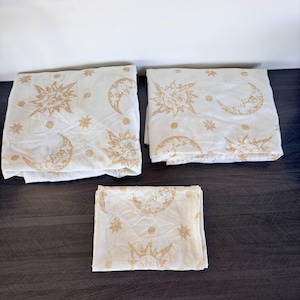 May include: Three folded white bed sheets with a gold celestial pattern of suns, moons, and stars. The sheets are arranged on a dark wooden surface. The celestial design is repeated on each sheet.
