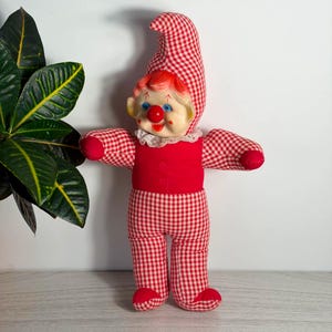 May include: A plush clown doll featuring a red and white gingham patterned hat, sleeves, and pants. The doll has a solid red torso, a white lace collar, and a molded plastic face with blue eyes, a bright red nose, and painted red cheek accents.