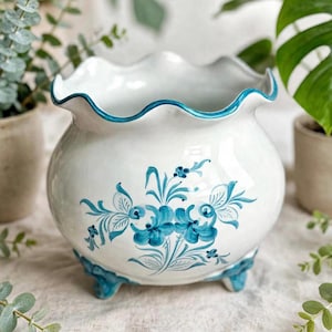 Könnte beinhalten: Ein weißer Keramikübertopf mit gewelltem Rand und drei kleinen Füßen. Die Außenseite ist mit einem handgemalten blauen Blumenmuster verziert, und der Rand ist mit einer passenden blauen Linie akzentuiert. Das Gefäß hat eine bauchige Form.