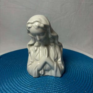 Puede incluir: Busto de cerámica blanca que representa a una mujer rezando, con las manos juntas. La estatua tiene el pelo suelto y está colocada sobre un fondo azul. La escultura muestra la cabeza y la parte superior del cuerpo.