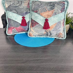 Peut inclure: Deux coussins décoratifs avec un motif dans les tons de gris, de rouge et d'orange. Chaque coussin présente un accent bleu clair et un pompon rouge. Les coussins sont placés sur une surface sombre.