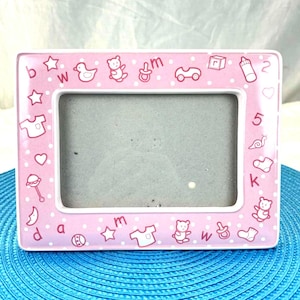 Peut inclure: Cadre photo rectangulaire rose pour bébé avec des pois blancs et des illustrations sur le thème des bébés. Le cadre présente des images d'un canard, d'un ours en peluche, d'une sucette, d'une voiture et d'un biberon. Le cadre est sur une surface bleue.