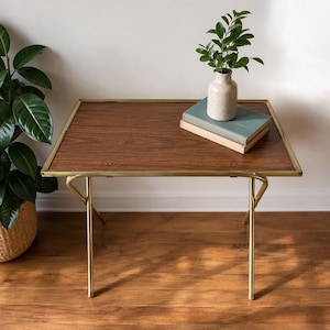 May include: A rectangular side table with a dark wood grain top and a thin gold-toned metal frame. The table features four slender legs and is topped with two stacked books and a small, light-coloured ceramic vase containing a green leafy plant.