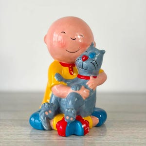 以下が含まれることがあります： ピーチ色の頭をした禿げたキャラクターの陶器製フィギュア。赤い襟の黄色いシャツを着て、赤い首輪と赤い鼻をした灰色の猫を抱えています。キャラクターは青いズボンと赤い靴を履いて座っている姿勢です。