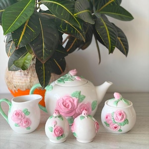 May include: A white ceramic tea set featuring raised pink rose blossoms and green leaves. The set includes a teapot with a lid, a creamer pitcher, a sugar bowl with a lid, and two small round shakers, all decorated with matching floral motifs.