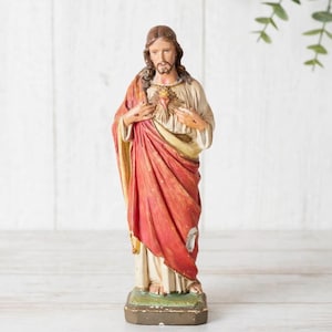 Könnte beinhalten: Eine stehende Figur mit langem braunem Haar und Bart, bekleidet mit einer cremefarbenen Tunika und einem drapierten roten Gewand. Die Hände liegen auf der Brust und zeigen ein stilisiertes Herz-Emblem, während die Figur auf einem Sockel steht.