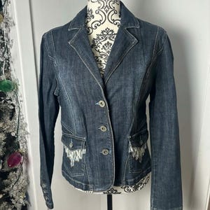 Peut inclure: Veste en jean bleu foncé de style blazer. Elle comporte trois boutons, deux poches avant avec broderies décoratives et des manches longues. La veste est présentée sur un mannequin.