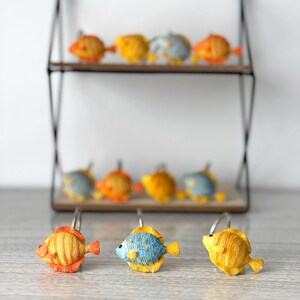 Puede incluir: Un conjunto de ganchos para cortinas de ducha con forma de pez de colores. Los ganchos son de varios tonos de naranja, amarillo y azul, con diseños detallados de aletas y escamas. Los ganchos se exhiben en una estantería decorativa.