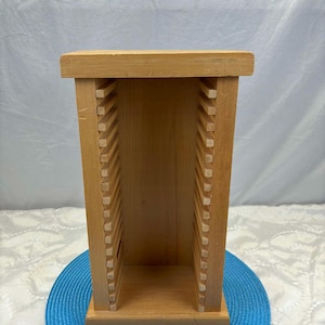 Może przedstawiać: Jasny, drewniany stojak do przechowywania płyt CD lub DVD. Stojak jest prostokątny z szeregiem pionowych przegród po każdej stronie. Góra i dół są z litego drewna. Stojak stoi na niebieskiej podkładce.