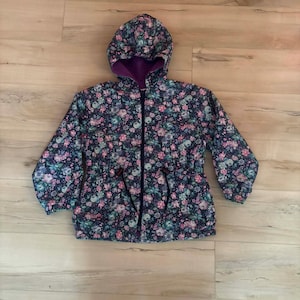 Puede incluir: Chaqueta con capucha con estampado floral en tonos rosa, azul y verde sobre fondo azul marino. La chaqueta tiene forro morado y cremallera completa. La cintura es ajustable con cordón.
