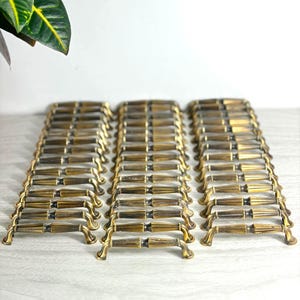 May include: A collection of numerous brass-toned metal drawer pulls arranged in three vertical rows. Each pull features a central tapered design with a dark, weathered finish and curved mounting ends, creating a uniform look for cabinet hardware.
