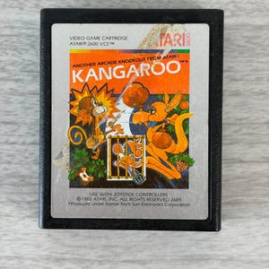 May include: A vintage Atari 2600 video game cartridge for the game "Kangaroo." The label features a colorful illustration of a kangaroo and a monkey, with the game title in large orange letters. The cartridge is black.