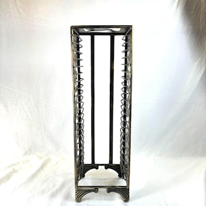 May include: A tall, rectangular metal rack with an aged bronze finish. The rack features vertical supports and a series of curved, decorative elements along the sides. The base has an ornate, scrolled design. The rack is designed to hold items.