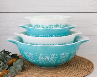 Puede incluir: Un juego de cuatro cuencos para mezclar apilables de varios tamaños, con un exterior de color turquesa brillante y un interior blanco. Cada cuenco está decorado con un patrón gráfico blanco que muestra figuras, plantas y globos aerostáticos.