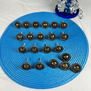 Op de afbeelding: Een verzameling antieke, bronskleurige kastknoppen met een touwachtige rand. De knoppen zijn gerangschikt op een blauwe placemat. Op de achtergrond staat een decoratief blauw en zilver ornament.