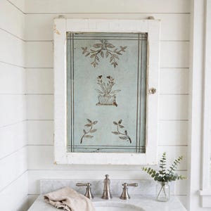 May include: A rectangular wall-mounted cabinet featuring a distressed white wooden frame and a central panel painted in a pale blue-green hue. The panel displays dark brown botanical illustrations of potted plants and leafy vines with thin border lines.