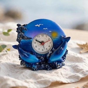 Op de afbeelding: Een ronde analoge klok met een blauwe oceaan-scène en twee gebeeldhouwde dolfijnen die uit de golven springen. De wijzerplaat is wit met zwarte cijfers en wijzers, tegen een achtergrond van een zonsondergang met drie witte vogels in de lucht.