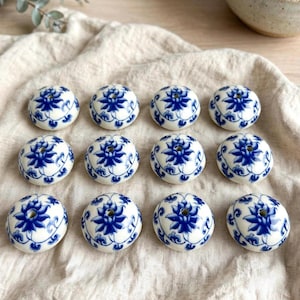 Könnte beinhalten: Zwölf runde Keramikperlen, angeordnet in drei Reihen zu je vier Stück. Jede Perle hat eine weiße Basis mit einem handgemalten blauen Blumenmuster und einem zentralen Loch. Die Perlen haben eine glatte, glänzende Oberfläche und eine einheitliche Form.