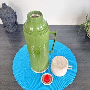 Peut inclure: Thermos vert vintage avec une poignée, une tasse blanche et un bouchon rouge. Le thermos est posé sur un tapis circulaire bleu. Le thermos a une surface texturée et une base blanche. La tasse a une poignée et le mot "Thermos" imprimé dessus.