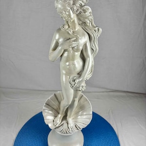 May include: A white statue of a woman, standing on a seashell base. The statue is approximately 19 inches tall. The woman has long flowing hair and is partially draped with fabric. The statue is a classic representation of a female figure.