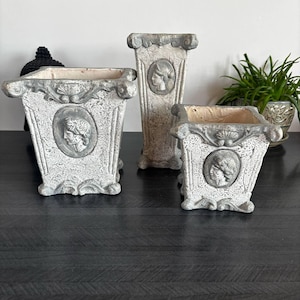 Può includere: Set di tre fioriere in stile antico di diverse dimensioni. Le fioriere sono bianco sporco con dettagli grigi e presentano un ritratto a rilievo classico. Sono realizzate in un materiale strutturato e sono adatte per uso interno o esterno.