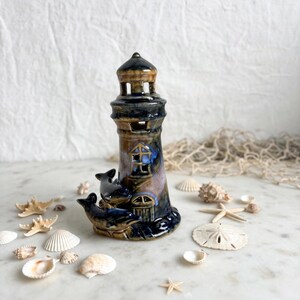 May include: A ceramic lighthouse figurine featuring a glossy glaze in shades of deep blue, brown, and tan. The structure includes cut-out windows and a door, with two sculpted dolphins attached to the base, surrounded by various seashells.