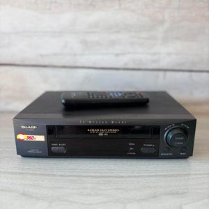 Puede incluir: Un reproductor de VHS estéreo Hi-Fi de 4 cabezales Sharp en color negro, con una etiqueta de zoom 360x y un mando a distancia negro a juego colocado encima. El panel frontal incluye una ranura para casetes, botones de encendido y expulsión.