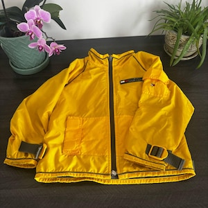 May include: A bright yellow jacket with a black zipper and trim. The jacket has a small pocket on the chest and two pockets on the front. The sleeves have adjustable straps. The jacket is on a dark surface.
