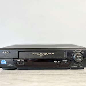 May include: A black Sharp VC-A582 VCR with the text "4-HEAD / SHARP SUPER PICTURE" and "VHS HQ". It features buttons for power, eject, play, stop, and channel selection. The VCR is on a light-coloured surface.