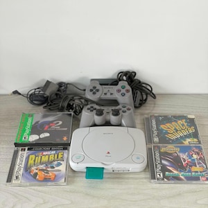 May include: A white PlayStation console with two gray controllers, power cables, and four game cases. The game cases include titles such as "Gran Turismo 2", "NASCAR Rumble", "Space Invaders", and "Digimon Digital Card Battle".