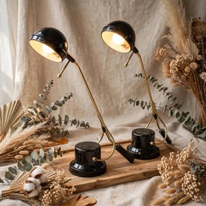 Peut inclure: Deux lampes de bureau avec des abat-jours en forme de dôme noir brillant et des bras en métal doré. Chaque lampe possède une base ronde noire avec un petit interrupteur et un col réglable. L'intérieur des abat-jours est blanc et lumineux.
