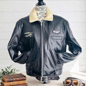 Puede incluir: Una chaqueta tipo aviador de cuero negro con cuello de piel sintética color crema, cremallera frontal plateada y cinturón con hebilla. El pecho presenta un parche bordado de un biplano a la derecha y un broche de ala de metal plateado a la izquierda.