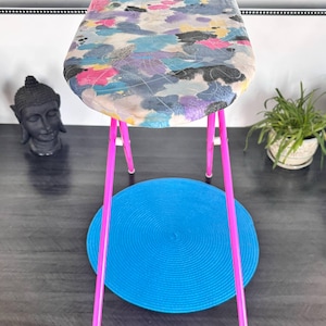 Puede incluir: Una tabla de planchar con una funda estampada con motivos florales coloridos y patas de color rosa brillante. Una alfombra redonda azul se encuentra debajo de la tabla. Una estatua de Buda negra y una planta en maceta están en el fondo.