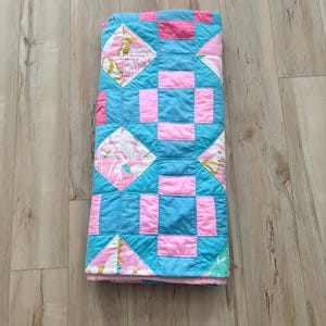 May include: Folded quilt featuring a patchwork design in shades of blue, pink, and white. The quilt has a repeating pattern of squares and diamonds. The quilt is folded neatly on a wooden surface.